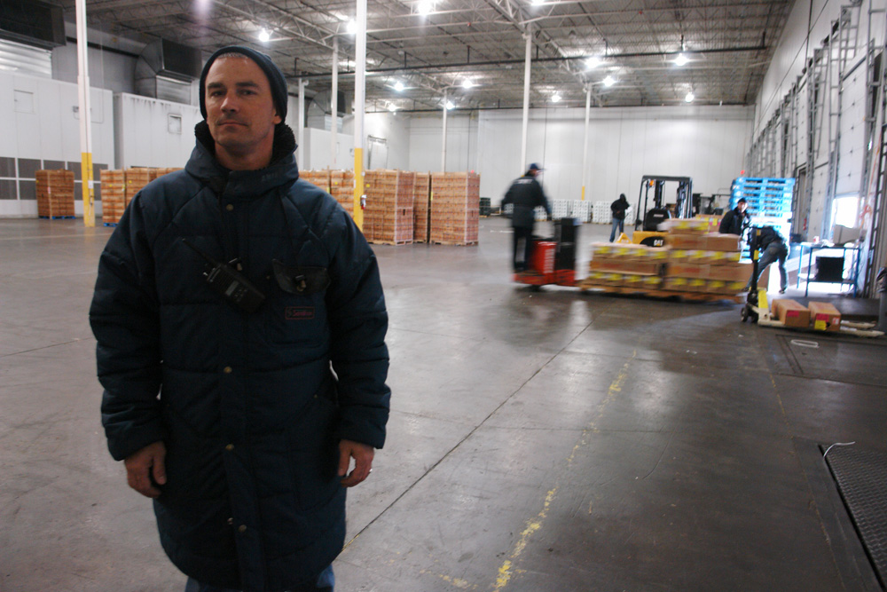 A floor supervisor in an insulated coat carrying two radios, crews moving pallets behind him