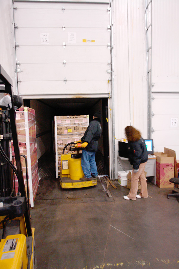A crew loading a trailer at Door 13 with an electric pallet jack, a dock terminal alongside